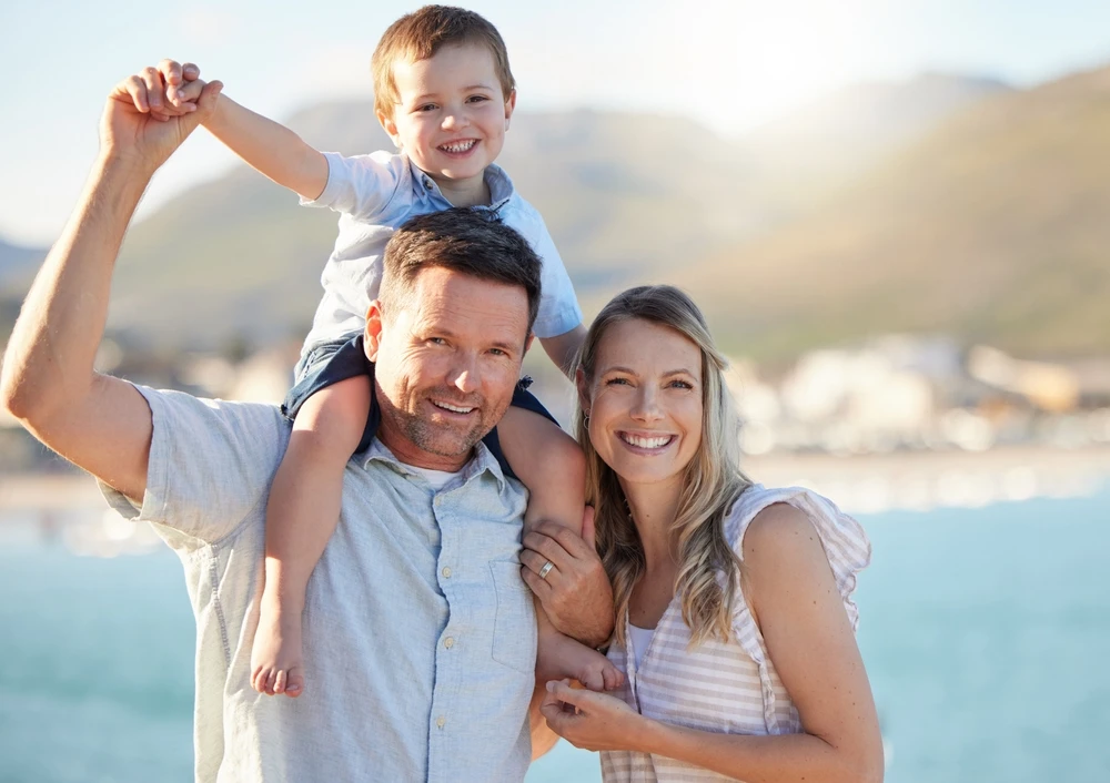 Smiling parents with a child sitting on his father’s shoulders by the water